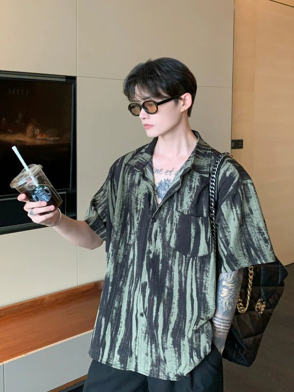 Person wearing a patterned shirt and holding a drink, standing indoors.